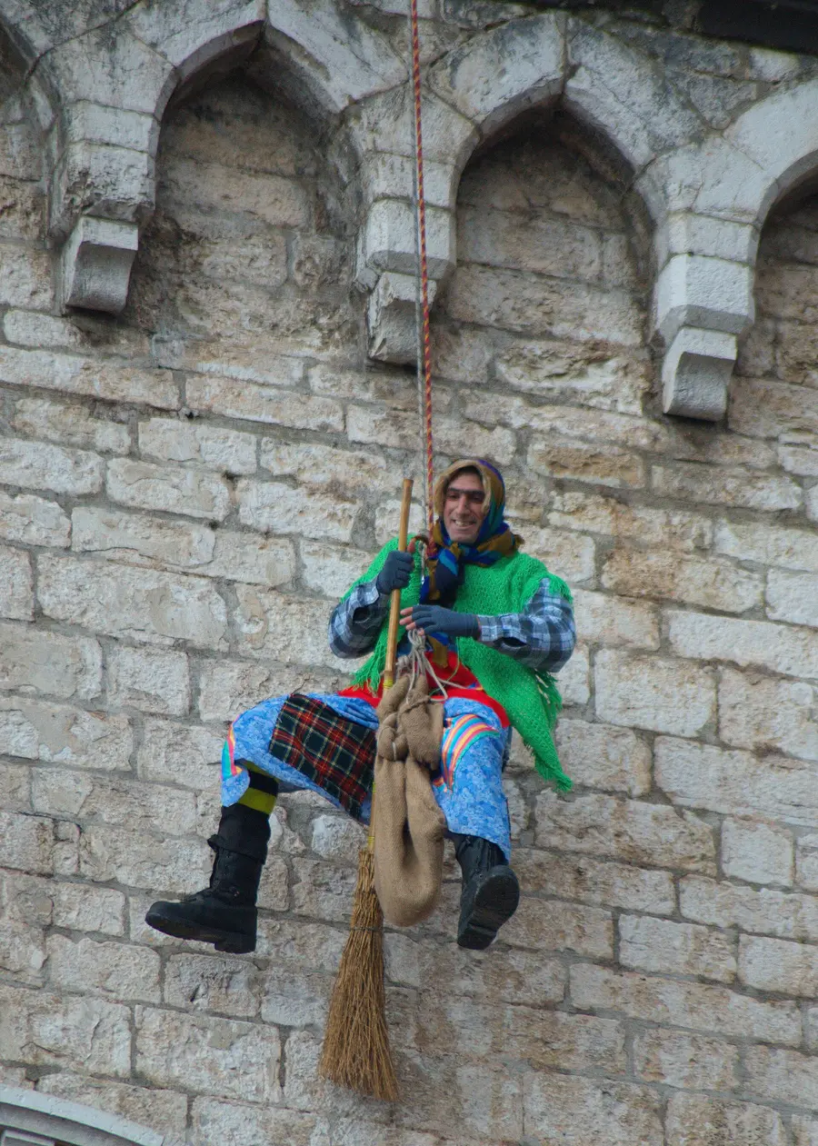 A woman dressed as La Befana with a broom and a sack descending a stone wall on a rope during an Epiphany festival in Gubbio, Italy