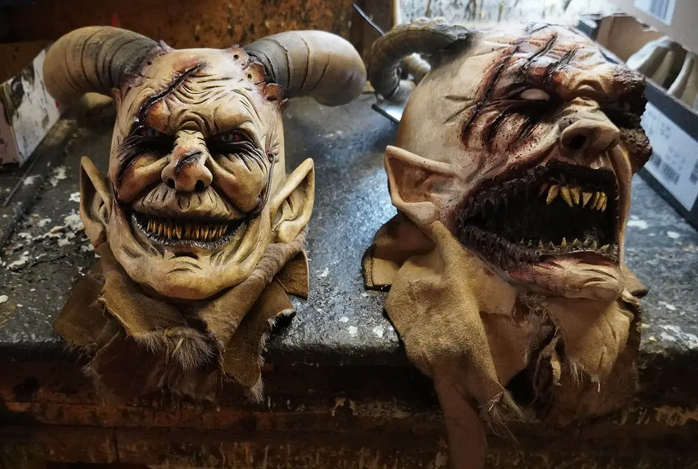 Two hand-made Krampus masks with horns, pointed ears and sharp teeth on a workshop bench