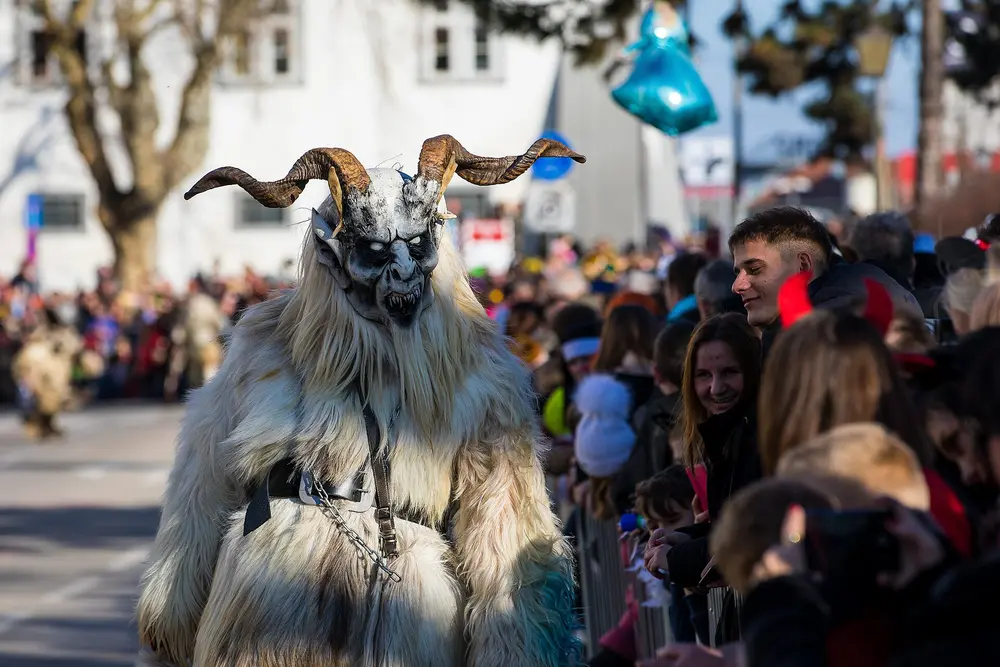 A white-furred Krampus with curled horns walking past a smiling crowd at a daytime Krampus parade