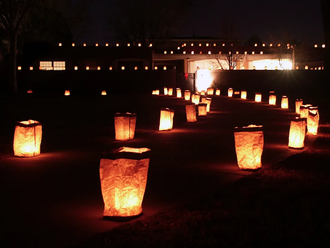 Rows of glowing paper-bag luminarias along a path and on the roofline of an adobe house at night