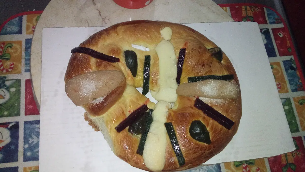 A Mexican rosca de reyes with candied fruit strips and a small white baby figurine on top