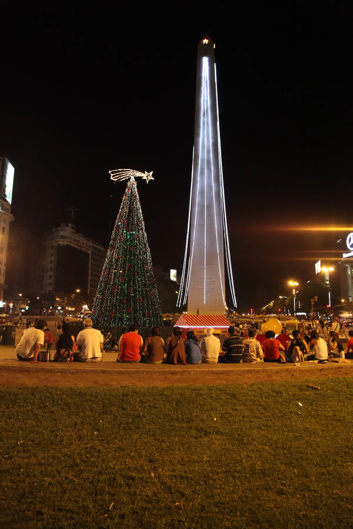 El Obelisco de Buenos Aires iluminado de noche junto al árbol de Navidad, con gente sentada en la plaza
