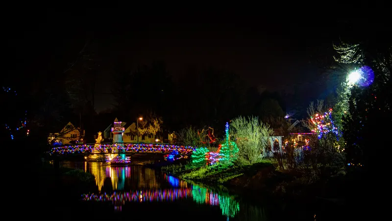 Lit footbridge and holiday village reflected in the river at Simcoe Christmas Panorama