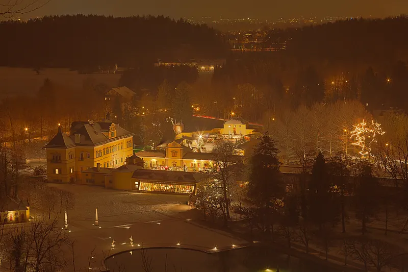 Blick über den beleuchteten Adventmarkt und den Park von Schloss Hellbrunn bei Nacht
